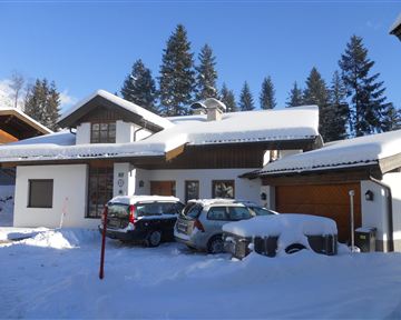 Ein modernes Haus in einer Schneelandschaft mit hohem Schnee. Im Hintergrund sind Nadelbäume und blauer Himmel zu sehen.
