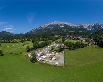 A vast, green landscape with mountains in the background. In the center, motorhomes and a small village are visible.