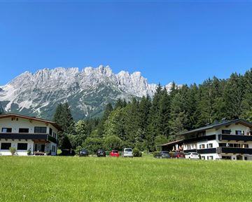 Ein schöner Blick auf zwei Gebäude inmitten von grünen Wiesen und einem Wald. Im Hintergrund sind beeindruckende Berge unter einem klaren blauen Himmel zu sehen.