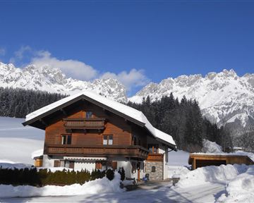 Ein charmantes Holzhaus im Schnee, umgeben von majestätischen Bergen. Der klare blaue Himmel lässt die winterliche Landschaft strahlen.