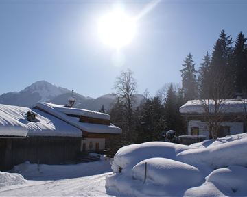Eine winterliche Landschaft mit schneebedeckten Dächern und sonnigem Himmel. Im Hintergrund sind Berge und hohe Bäume zu sehen.
