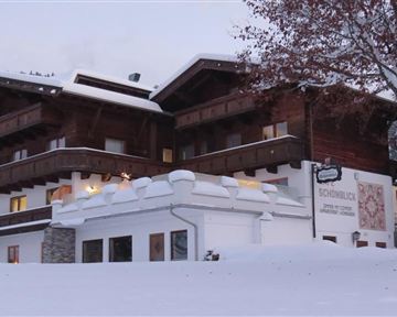 Ein gemütliches Holzhaus im Schnee, umgeben von einer winterlichen Landschaft. Die Fassade ist traditionell mit Balkonen und großen Fenstern gestaltet.
