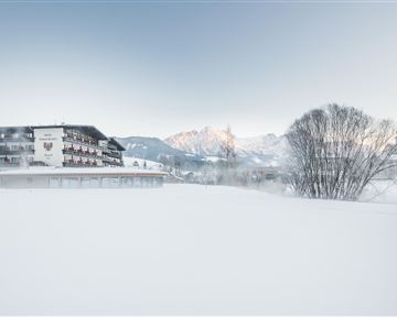 A snow-covered landscape with a hotel in the foreground and mountains in the background. The sky is clear and the scene radiates calmness and cold.