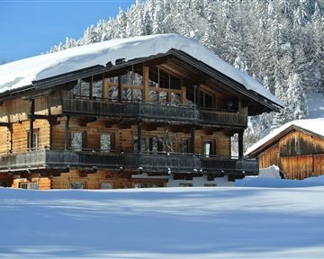 Ein Holzhaus im Schnee mit einem Dach voller Schnee. Im Hintergrund sind verschneite Bäume zu sehen.