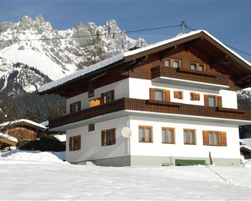 Ein schönes, zweigeschossiges Chalet in den Alpen, umgeben von Schnee. Im Hintergrund erheben sich majestätische Berge unter einem klaren Himmel.