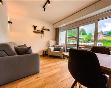 Ein modernes Wohnzimmer mit einem grauen Sofa und einem Esstisch. Große Fenster bieten einen Blick ins Grüne.